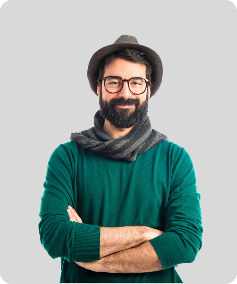 A smiling man with a beard, glasses, & a hat, wearing a green sweater & a scarf, with arms crossed against a gray background.